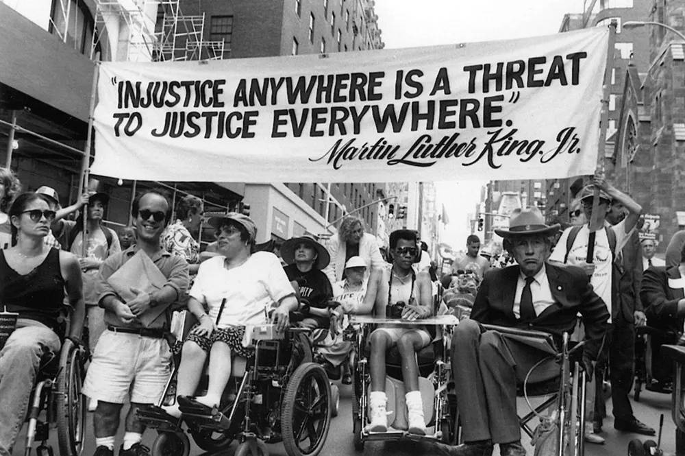 People in wheelchairs participate in a street rally holding a banner with a quote.